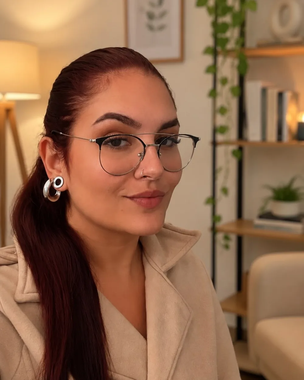 Rafaella Maldonado de óculos e casaco bege, em um ambiente aconchegante com plantas e uma estante de livros ao fundo